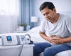 Middle-aged Indian man sitting on a bed with stomach discomfort beside a CPAP machine in a blue-toned bedroom.