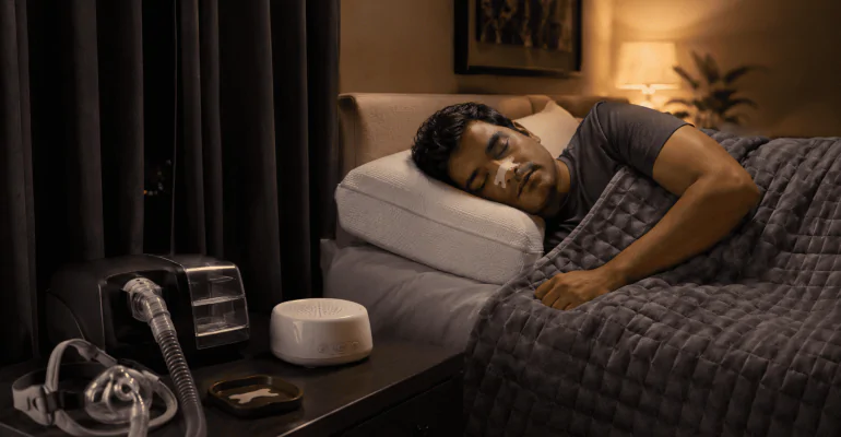 Person sleeping peacefully in a bedroom with CPAP machine, weighted blanket, nasal strip, blackout curtains, and white noise machine.
