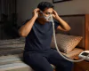Indian man adjusting a CPAP mask before sleep with a CPAP machine on the bedside table.