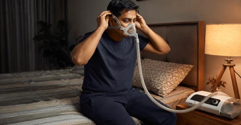 Indian man adjusting a CPAP mask before sleep with a CPAP machine on the bedside table.